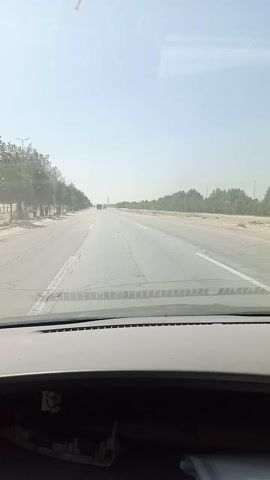 This is a video taken from inside a vehicle, looking out the windshield at a road. The road appears to be an airport runway or a similar long, paved stretch of ground. The time of day is daytime, indicated by the bright, clear sky. The weather appears to be clear and sunny, with a slight haze in the distance. 

The scene is a flat, open landscape with sparse vegetation on either side of the road. The vegetation consists of trees and shrubs, suggesting a dry or arid climate, consistent with the location of Dhahran, Saudi Arabia. The road itself is marked with white lines, and there are some cracks and imperfections on the surface, indicating it has been used for some time. In the distance, there are a few structures and possibly vehicles, but they are not clearly discernible due to the haze and distance.

The video captures a sense of forward motion, as the road stretches out into the horizon. There are no people visible in the frame. The overall mood is neutral, focusing on the journey and the environment. There is no visible text in the video.