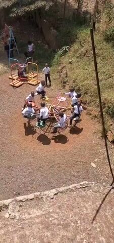 The video shows a playground area with several children in white shirts and dark pants playing on various equipment. In one area, a group of children is on a merry-go-round, some sitting and others pushing it. The camera pans to a trampoline where several children are jumping, lying down, and interacting. Adjacent to the trampoline is a slide, swings, and other playground structures. A building with a red roof and a painting on the wall is visible in the background. The playground is on a dry, grassy surface with trees and bushes surrounding it.