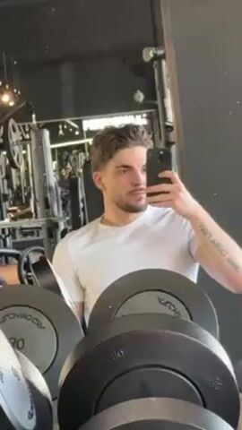 A young, light-skinned man with brown hair and a stubbly beard stands in front of a mirror in a gym, taking a selfie. He is wearing a white t-shirt. Behind him are rows of weights, including 30-kilogram dumbbells. In the background, gym equipment is visible, and another person is working out. The gym has dark walls. The man holds his phone in his right hand, and a tattoo is visible on his forearm. He glances towards the side as he takes the photo.