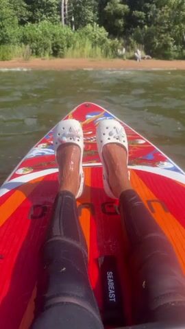 The video shows a first-person perspective of someone sitting on a paddleboard in the water. The person's legs are extended forward, and they are wearing black leggings and white Crocs decorated with charms. The paddleboard is red with colorful graphics. In the background, there is a shoreline with trees and a body of water. The sun is shining, indicating it is daytime.