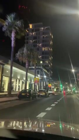 This video shows a nighttime view from inside a vehicle moving along a street. The camera is pointed forward and slightly upwards, capturing the illuminated facade of a tall, modern building on the right side of the street. The building has numerous balconies with curved edges and is lit with warm, interior lights. A sign on the building reads "MR. ZAVOS GROUP" in green neon letters. Below the tall building, there are lower structures with storefronts, one of which has "CARA" in illuminated lettering. Several cars are visible on the road, including a black car in the foreground on the left, a gold SUV in the middle of the road, and a white SUV in front of it. Traffic lights appear to be green. Palm trees with illuminated trunks are visible on the left. The street is paved, and there are white dashed lines marking lanes. The overall environment is urban and appears to be a commercial or residential area.