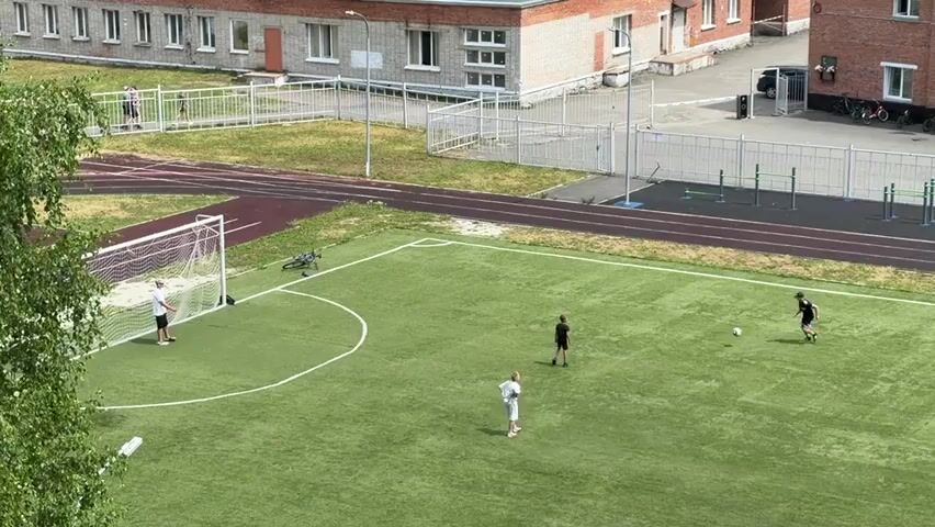 A high-angle shot shows four young people on a green artificial turf soccer field. Two of them are playing soccer with a ball, while one is standing near a soccer goal and another is kneeling on the ground. In the background, a brick building with white trim and windows is visible, along with a fenced-off track and field area. The sky is overcast.