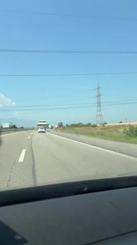 The video shows a view from inside a car driving on a highway on a clear day. The highway is paved and has lane markings. There are other vehicles on the highway, including a large truck towing a white yacht. A white car with flashing orange lights, likely a support vehicle, is in front of the yacht. In the background, there are fields, trees, and power lines. The sky is blue.