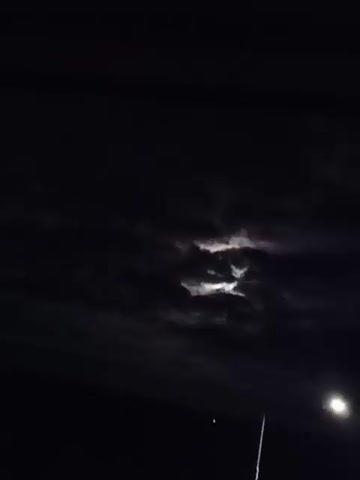 A nighttime shot shows the moon partially obscured by dark clouds. A bright streetlamp is visible in the lower right corner. Two thin, vertical poles are also in the frame, one in the center and one to the right. In the distance, some vegetation and a road are visible.