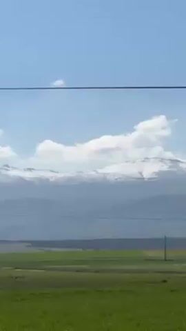 The video shows a landscape with green fields, telephone poles, and mountains in the background. The mountains have snow on their peaks. The sky is blue with some clouds. The video appears to be taken from a moving vehicle, as the landscape shifts in the frame.