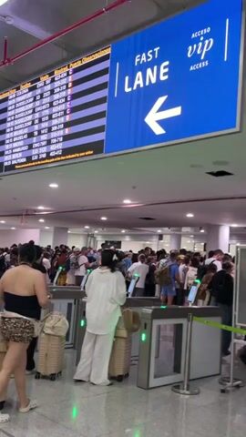 The video shows a busy airport departure board displaying flight information with destinations including Montreal, Dusseldorf, and Berlin. Below the board, a long queue of people is waiting to go through security gates. The gates have green lights indicating they are open. Many people are carrying luggage. The overall atmosphere appears to be one of congestion and waiting.