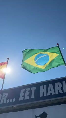 The video shows several national flags waving in the wind against a bright blue sky. In the foreground, a sign with Dutch text reads "GEEF HET DOOR..." which translates to "PASS IT ON...". The sun is shining brightly, creating a lens flare and backlighting the flags. The flags visible are the Brazilian flag, the Spanish flag, and the Jamaican flag. There are also hints of other flags behind the main ones. The scene appears to be outdoors, possibly at an event or a business with a celebratory atmosphere.