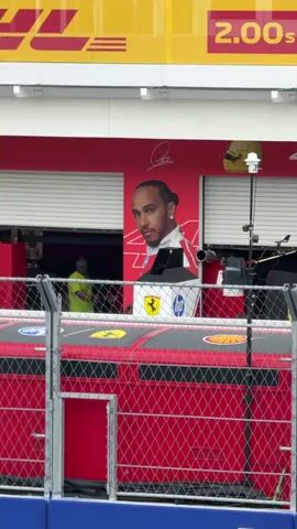 The camera pans out to reveal a Formula 1 pit lane and surrounding area. The pit lane is bordered by a fence and a large sign for crypto.com. Above the pit lane is a series of balconies with people watching. On the top level, the Hard Rock Stadium is visible. The pit lane itself has advertising for DHL, Ferrari, and HP. There are large portraits of Lewis Hamilton and Charles Leclerc on the pit garages. Several people can be seen working in the pit garages.