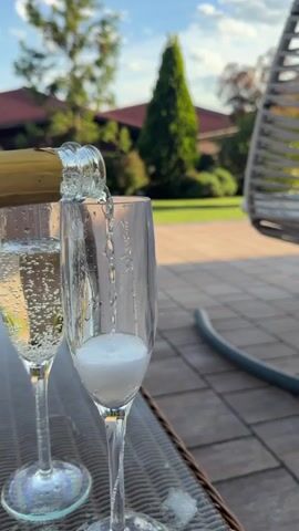 A person pours a sparkling beverage from a glass bottle into a flute glass. The glass fills with a bubbling liquid that quickly produces a thick white foam at the top. Another glass of the same beverage, filled with a similar amount of liquid and bubbles, stands beside it. The glasses are placed on a table with a dark, textured surface that resembles tile or brick. In the background, there is a green lawn, trees, and houses with red roofs under a partially cloudy sky. To the side, a portion of a gray hanging chair is visible. A length of gray tubing lies on the ground. The overall lighting suggests it is daytime.