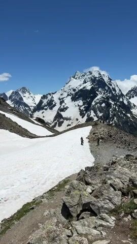 The video shows a scenic mountain landscape with snow-covered peaks under a clear blue sky. The camera pans across the terrain, revealing rocky outcrops in the foreground and a winding path. Two hikers are visible on the path, walking along a snow-covered ridge. In the background, majestic, snow-capped mountains dominate the view, with patches of green vegetation visible on some slopes. The overall impression is one of a vast, natural, and mountainous environment.