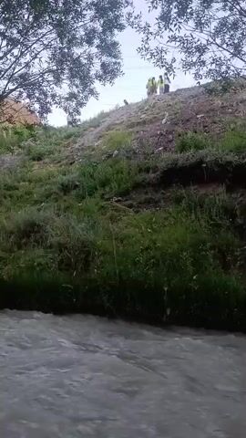 A medium-angle shot shows a rushing river in the foreground, with a grassy slope rising behind it. Several people are visible on top of the slope, silhouetted against the sky. Trees frame the top of the shot. The scene appears to be outdoors, possibly in a natural setting like a park or along a riverbank. The lighting suggests it is daytime, with a bright sky. The overall atmosphere seems relaxed and casual, with people enjoying the outdoors. No visible text is present.