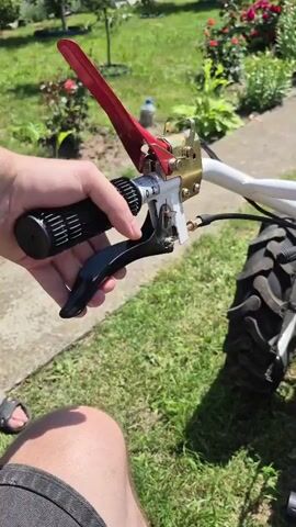 A hand grips the handle of a tiller, the red lever likely a throttle or clutch. The tiller has two large tractor-like wheels and a red engine casing. The background is a grassy yard with flower beds. The video pans across the tiller, showing the engine and wheels.