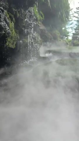 The video shows a close-up, low-angle view of a small waterfall cascading over moss-covered rocks. Dense white mist or fog hovers close to the ground, obscuring the base of the waterfall and swirling around the rocks and surrounding vegetation. The mist creates a serene and mystical atmosphere. In the background, a lush green cliff face is visible, with ferns and other plants growing on it. Sunlight filters through the foliage, creating dappled light and highlights. The overall impression is one of a tranquil, natural setting with an emphasis on the interplay of water, mist, and greenery.