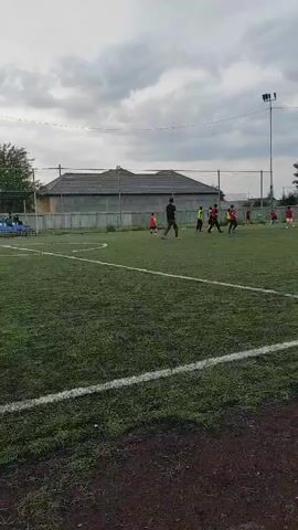 A group of children are playing soccer on a cloudy day on a green artificial turf field with white lines. There are buildings and trees in the background, and a tall light pole.