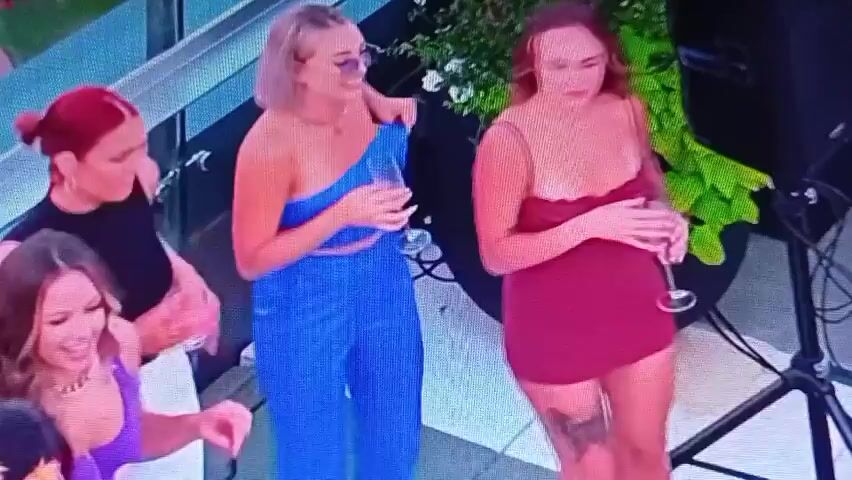 The video shows a group of five women at an outdoor gathering, possibly a party or event. One woman in a blue strapless top and matching pants is adjusting her clothing. To her right, another woman in a red dress is also adjusting her attire. To the left of the woman in blue, a woman with red hair is holding a drink and observing. In the foreground, a woman in a purple top is smiling and looking down. In the background, a woman with long dark hair wearing headphones is seen at a DJ setup. The environment appears to be a rooftop or elevated area with a view of the sky, and some plants are visible.