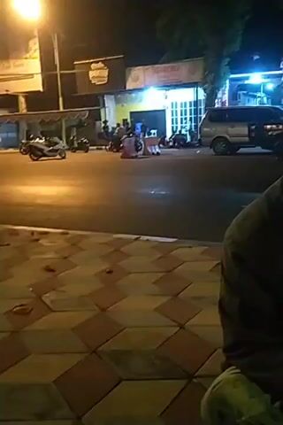 The video shows a street scene at night. There is a road with vehicles, including cars and motorcycles. Several people are gathered on the sidewalk and near the road. Buildings with lights are visible in the background. The overall impression is of a public area with activity at night. The camera is handheld and the footage is somewhat shaky.