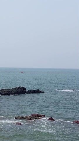 The scene is a wide shot of the ocean, captured from a high vantage point. The sky is overcast. The water is a muted blue, with white foam forming around rocky outcrops in the foreground. In the distance, a small white boat with figures visible on board is moving across the water. The overall atmosphere is serene, despite the somewhat bleak weather.