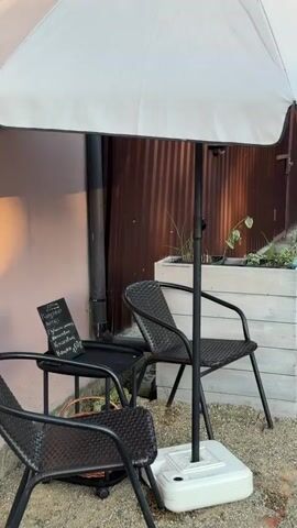 The video shows an outdoor seating area with two black metal chairs and a table under a large white umbrella. A white planter box with plants sits behind the chairs. A pink wall is visible in the background with a sign that says "ENGLISH LANGUAGE" and a phone number. To the left, a black metal archway stands next to a doorway draped with white macrame curtains. A vertical sign with Russian text that reads "COFFEE TO GO" is attached to the wall next to the archway. Sunlight casts shadows on the wall.