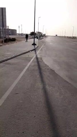 The video shows a long, straight asphalt road with a solid white line marking the edge of the lane. A person in dark clothing is walking away from the camera on a paved sidewalk to the left of the road. There are several evenly spaced lampposts along the roadside. A bus shelter is visible further down the road. Some buildings and a communication tower are visible in the background.  The sky is bright and hazy. The camera appears to be moving slowly along the road.
