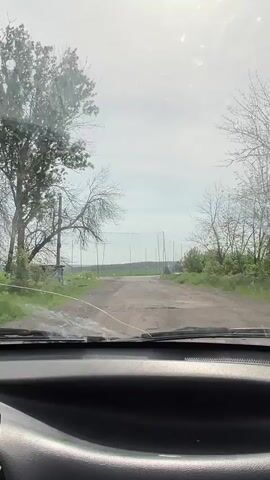 The video shows a view from inside a car, looking out through the windshield as it drives along a dirt road. The road is somewhat cracked and uneven. On either side of the road are trees, mostly bare, with some green foliage visible. In the distance, the road passes under a series of vertical poles with hanging cords, arranged to form a sort of netting or screen. The sky is overcast with bright sunlight behind the clouds. The car's dashboard is visible at the bottom of the frame. The camera is relatively stable, with some minor vibrations due to the road condition.