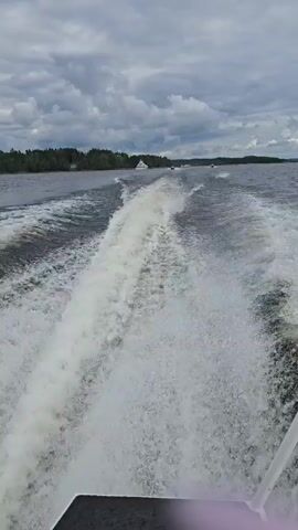 A POV shot from the back of a moving boat shows a large wake trailing behind. The water is choppy, and the sky is overcast with clouds. In the distance, other boats can be seen, as well as a tree-lined shore. The scene is a lake or a large body of water, likely on a cloudy day.