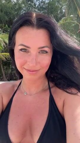 A woman stands on a beach and smiles at the camera. She is wearing a black bikini top and a silver necklace. The woman has dark hair and a fair complexion with some freckles. The beach is sandy with palm trees in the background. There are a few people walking in the background.