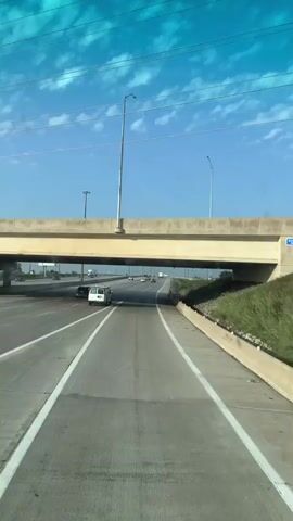 A first-person perspective video captures a vehicle traveling on a highway. The vehicle passes under an overpass with multiple lanes of traffic visible on both levels. Several other vehicles, including cars and a white van, are also seen on the highway. The sky is blue with scattered clouds, and the surrounding environment consists of grassy areas and a concrete barrier. The video appears to be filmed during daylight hours.