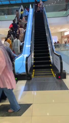 The video shows a busy mall escalator with many people going up. The escalator has black steps and a blue illuminated side. There are signs on the sides of the escalator. People are dressed in casual and traditional clothing. Some people are looking at their phones. The mall appears to be modern with a glass ceiling and storefronts visible in the background.