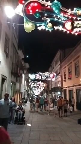 A low-angle shot looking up at a narrow, cobblestone street at night, lined with buildings on either side. The street is illuminated by strings of colorful Christmas lights that are draped across the overhead space. Several people are walking down the street, some appearing to be tourists. In the foreground, a woman in a grey t-shirt and jeans walks towards the camera, while beside her, a person in a wheelchair is being pushed by someone unseen. Further down the street, more people are visible, along with more festive decorations hanging between the buildings. The overall atmosphere suggests a lively evening in a European city.