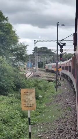 A train is traveling alongside a green, overgrown area. There is a signpost in the foreground with text in both English and what appears to be a non-English script, and the English text reads "FOR CALLING ON SIGNAL STOP AHEAD OF THIS BOARD". There are railway tracks and signals in the background, and another train is visible further down the tracks. The sky is overcast with clouds.
