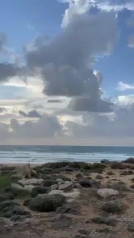 The video shows a view of the sea with waves rolling in under a cloudy sky. A patch of bright rainbow colors is visible through a break in the clouds. The foreground is a sandy area with low-lying vegetation and some rocks. The camera zooms in slightly to focus on the rainbow effect in the clouds.