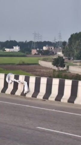 A roadside view captures a rural landscape with houses, fields, and power lines under a cloudy sky. A highway barrier with black and white stripes runs along the foreground, with a road to its left. The scene is captured from a moving vehicle.