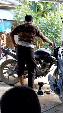 From a low angle, a person wearing a brown shirt and black pants stands behind a black motorcycle. They have their left hand on their hip and their right hand on the handlebar. A drill and some other tools lie on the ground next to the motorcycle. The setting appears to be an outdoor workshop. The camera angle shifts slightly to the left, then back to the initial position. Another person is visible in the foreground, with only the back of their head in view. A sign is visible in the background. The motorcycle appears to be undergoing repairs.