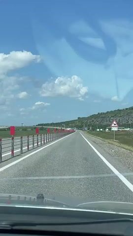 A first-person perspective video shows a car driving on a highway on a sunny day. The highway is paved and has two lanes in each direction, separated by a white line. On the left side of the road, there is a metal barrier with red and white reflectors. In the distance, there are green hills and a blue sky with white clouds. A few trucks and other vehicles are visible on the highway. There are also some signs on the side of the road, including a triangular warning sign with an image of an animal. The car's dashboard is partially visible in the foreground. The video is brief and does not contain any dialogue or music.