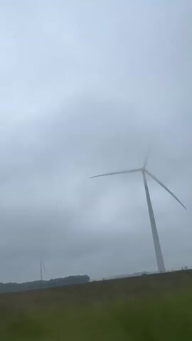 This media file depicts a foggy landscape in Rimšai, Lithuania. The primary subject is a tall, white wind turbine with three blades, partially obscured by thick, gray clouds. The turbine stands on a flat plain, with a line of dark trees or foliage visible in the distance. The foreground consists of a grassy field, also appearing somewhat muted due to the weather. Another, more distant wind turbine is faintly visible to the left, also shrouded in mist.

The weather is overcast and very foggy, creating a somewhat somber or ethereal atmosphere. The light suggests it is daytime, but the fog limits visibility and softens the overall scene. There is no discernible activity beyond the implied operation of the wind turbine, which is likely generating power. No people are visible in the media. The setting is clearly rural, characterized by open land and wind energy infrastructure. There is no visible text in the file.