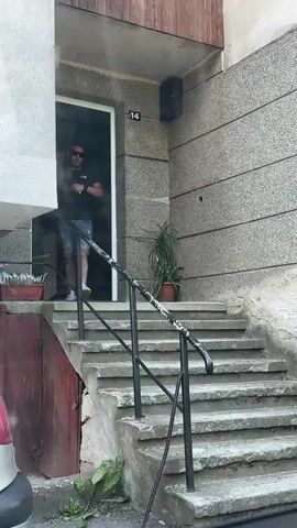 A man wearing a black t-shirt, denim shorts, sunglasses, and white sneakers stands on a concrete stairway outside a building. He is smiling and holding a small object in his right hand. The stairway has a metal railing. The building's facade is made of textured concrete and wood paneling. A potted plant is visible to the right of the doorway. The number 14 is displayed on the building above the doorway.