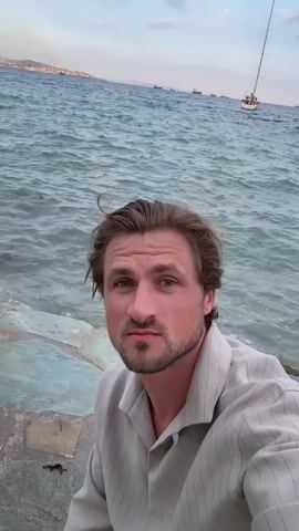 A man with brown hair and a beard looks into the camera. He is wearing a tan collared shirt with thin vertical stripes. The man is sitting on large, flat stones near the ocean. Waves are crashing against the shore. In the background, there are boats on the water and a coastline with buildings. The sky is cloudy. The man makes a pained expression at 00:03.