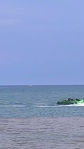 A bright green, car-shaped boat is cruising across calm, blue water under a pale blue sky. The boat is leaving a white wake behind it. In the distance, there are faint outlines of land and a small boat. The foreground shows the choppy surface of the water closer to the shore.