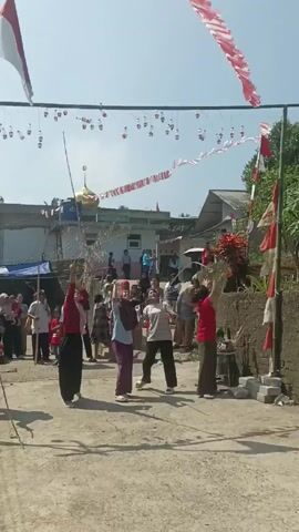 A group of women play a game outdoors in a village, jumping to reach dangling objects tied to a string strung between two poles decorated with Indonesian flags. The women are of different ages and wear casual clothing. The game appears to be part of a community celebration, with many other people watching in the background.  There is a building with a golden dome behind the group and houses and vegetation. The sky is clear and sunny.