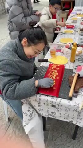 A woman in a gray jacket and blue jeans is seated at a table outside, focused on applying gold detailing to a red rectangular object. She is wearing gloves. The table is covered with a patterned white tablecloth. To her right are an axe-shaped object, a decorative brown block, and a bottle. In the background, a boy in a white jacket and glasses is also working on a craft project at the same table. Other people walk by in the background. The setting appears to be an outdoor market or craft fair.