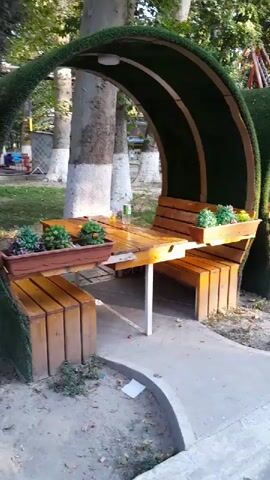 The video pans across a row of outdoor seating areas, each with a semicircular, arched canopy covered in artificial turf. The canopies are supported by wooden structures and feature built-in wooden benches and tables. Small planter boxes with greenery are mounted on the tables. The seating areas are arranged along a paved walkway, with trees and foliage visible in the background. The ground around the seating areas is covered with dirt and some scattered leaves. In the far background, a playground structure can be seen. The lighting suggests it is daytime.