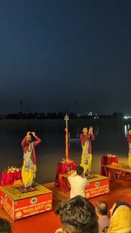 This video captures a religious ceremony, likely a "Maha Aarti," taking place at dusk or night by a body of water. Three men in traditional Indian attire, consisting of maroon kurtas and yellow dhotis with blue and yellow sashes, stand on elevated platforms. They are performing a ritual, with their hands clasped together in prayer or greeting. Each platform has decorative cloths and several small lit lamps (diyas) placed around the edges. A trident-like object, possibly representing Lord Shiva, is prominently displayed on a stand behind the central performer.

In the foreground, a crowd of spectators is gathered, with some individuals also seen with their hands in prayer. The scene is set against a dark sky, with distant lights visible on the opposite shore of the water, suggesting an urban or semi-urban location. The water itself reflects some of the ambient light. The overall atmosphere appears to be serene and devotional.

Visible text on the orange boxes beneath the performers' platforms reads: "SHREE RAGHUNATH JI CORRIDOR VIKAS COUNCIL, JAMMU" and "MAHA AARTI" along with "TOURISM FEDERATION JAMMU." This text clearly indicates the location as Jammu, India, and identifies the event as a "Maha Aarti" organized by the Shree Raghunath Ji Corridor Vikas Council and the Tourism Federation. The time of day is evident from the deep twilight or nighttime sky.
