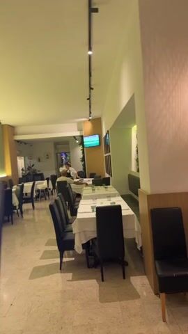The video pans across a restaurant interior. The restaurant is clean and modern, with white tablecloths on the tables and black chairs. There is a television mounted on the wall, showing a soccer game. Several tables are set for dining, with glasses and cutlery in place. A person walks into the frame from the right side and moves out of focus. Another person, wearing a white shirt, is seated at a table in the background. The lighting is dim, with spotlights illuminating the tables and the television. Plants are visible on the wall behind the television.