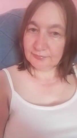 The media file is a close-up selfie of a middle-aged woman with brown hair and fair skin. She is wearing a white tank top and appears to be indoors, possibly in a living room or bedroom, given the soft lighting and the presence of a blue patterned fabric in the background, likely upholstery. Her expression is neutral, with a slight smile. The time of day is not explicitly indicated, but the lighting suggests it could be daytime. There is no visible text in the media file. The location context provided is Berezynka, Ukraine.
