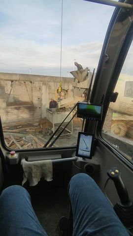 From inside a crane cabin, a view of a construction site is visible through the windshield. Workers are present, and a concrete mixer is in operation. A partially constructed brick building with wooden supports is also visible.
