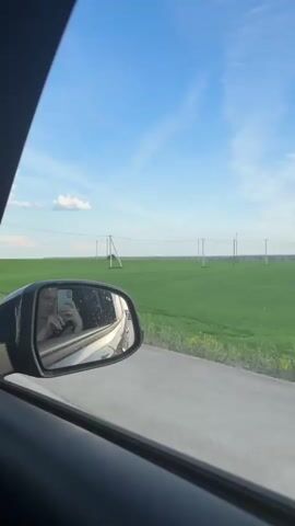 The video, shot from inside a moving car, captures a young woman's reflection in the side mirror. She holds a smartphone, taking a selfie or recording herself. The car is on a road next to a vast green field under a blue sky with some scattered clouds. In the distance, there are several utility poles with power lines stretching across the field.