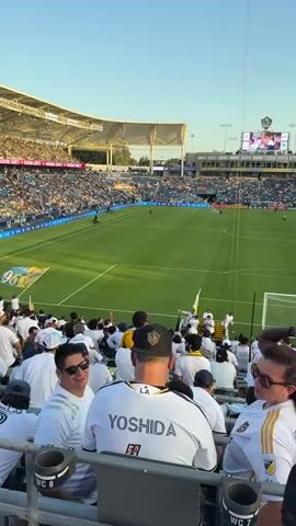 The video shows a soccer game from the perspective of a spectator in the stands. The field is visible with players, and the stands are filled with people wearing white shirts, many with player names on the back. The sky is clear and blue, and the sun is shining. In the background, a large screen displays graphics. The video pans across the stadium and the crowd.