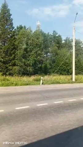 The video shows a roadside view from a moving vehicle. On the left side of the road, there is a white utility pole with a cell tower in the background. A speed limit sign with "50" is visible on the right side of the road. A pedestrian crossing sign is also visible on the left, followed by a crosswalk. The road is paved and has white lane markings. Beyond the road, there are trees and vegetation. A white car is seen driving in the distance on the right side of the road. The sky is blue with scattered clouds.
