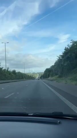 A view from inside a vehicle driving on a highway. The highway has multiple lanes and is bordered by lush green trees and vegetation on either side. The sky is partly cloudy with a mix of blue sky and white clouds. Several other vehicles, including cars and a truck, are visible on the road ahead. The road appears to be winding and goes uphill in the distance. There are streetlights and signs along the highway.