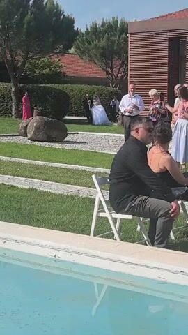 A person is filming an outdoor wedding ceremony. A bride in a white wedding gown and veil walks down a stone path on a grassy lawn, escorted by a man in a black suit who holds her arm and a bouquet of white flowers. Several guests are visible in the background, some standing and some seated. In the foreground, a man in a black shirt and a woman in a black top are seated in white folding chairs next to a light blue swimming pool. The setting is a sunny, well-maintained garden with green grass, trees, and a modern building with horizontal wood paneling.